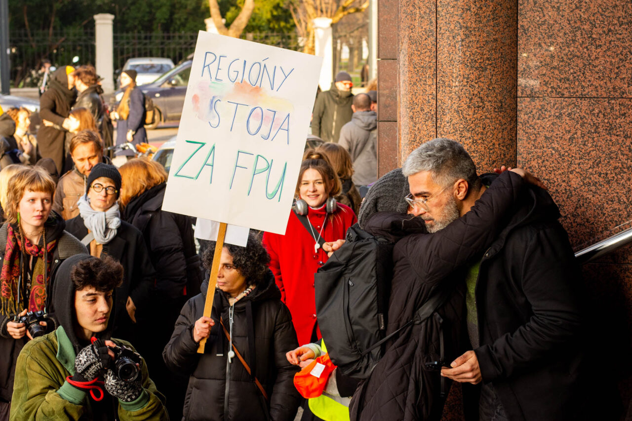 Fotografia skupiny ľudí z protestu pred budovou Fondu na podporu menia.  Jedna z účastníčok drží transparent s nápisom "Regióny stoja za FPU".