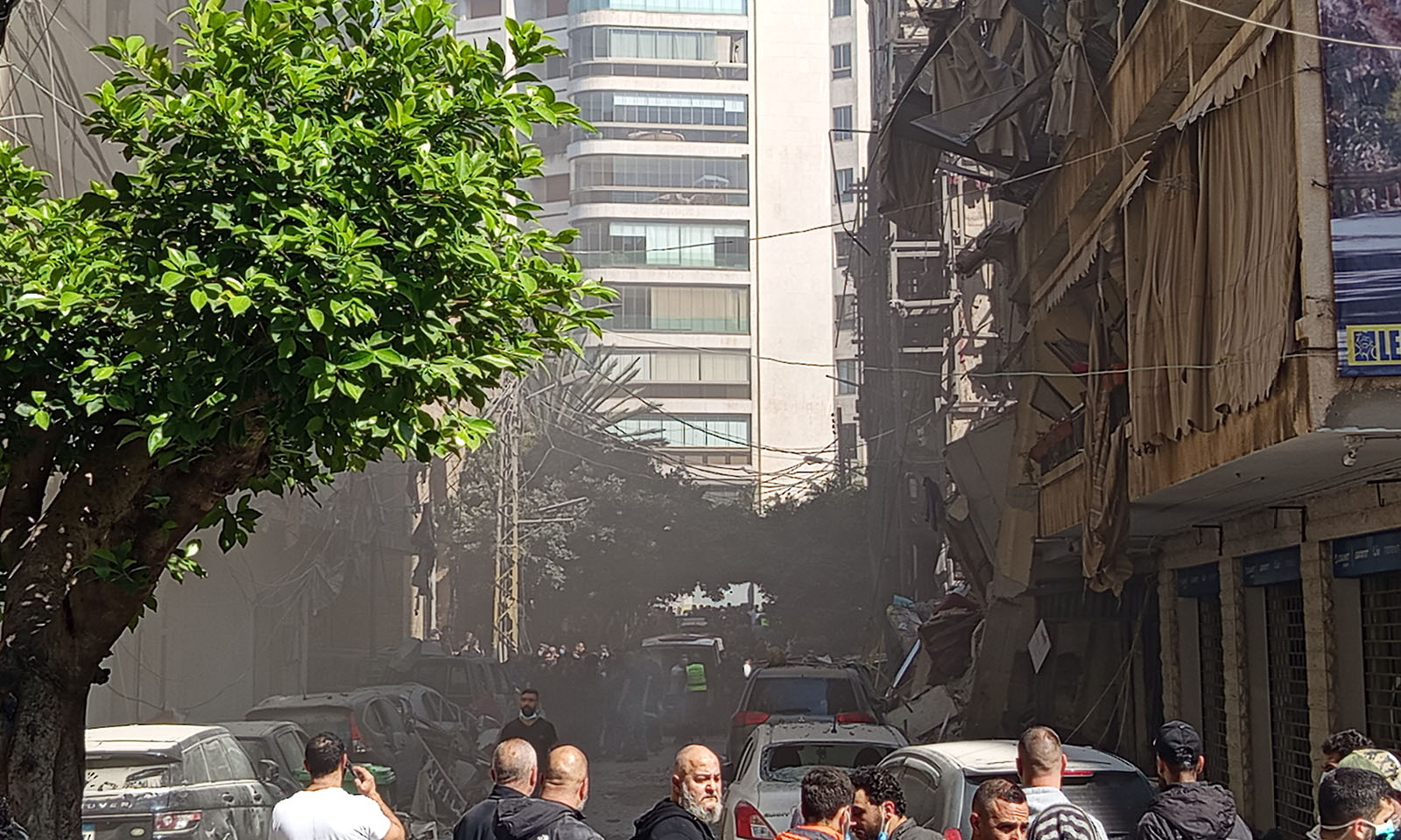 Ulica so zbombardovanými budovami. Autá pokryté prachom. Skupina ľudí stojí v bezpečnej vzdialenosti.